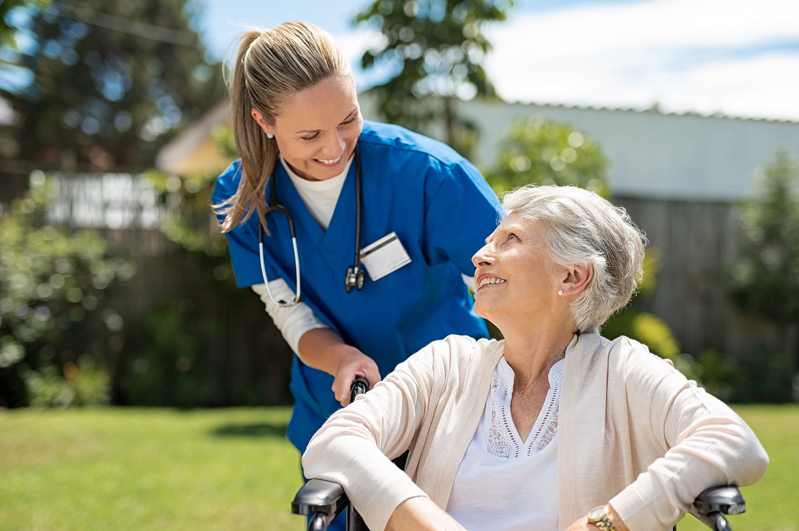Caregiver smiling with an older woman in a wheelchair outdoors.